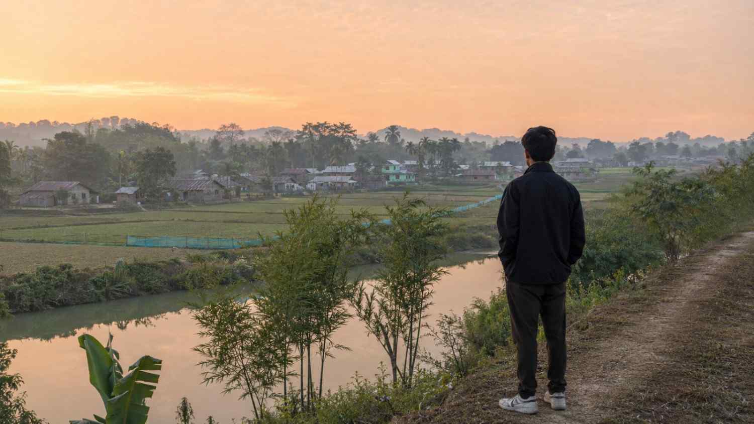 Young person looking across an Assamese river landscape symbolising memory, belonging and connection with the homeland.